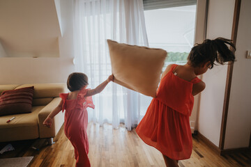 Wall Mural - Two young girls in colorful dresses playfully engage in a pillow fight in a sunlit living room, capturing the joy and energy of childhood fun.