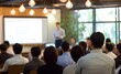© 为轩 张 - Asian business training presentation in a modern office, audience seated and listening to the speaker with light bulb pendant lights, whiteboard, and laptop displaying PowerPoint content.