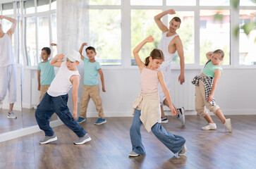 Focused preteen girl performing dynamic hip hop moves in group class for kids, led by skilled young male dance instructor in vibrant urban studio