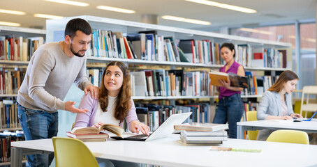 Canvas Print - Young positive interested couple of students spending time together in library, reading books and browsing educational websites on laptop