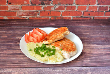 Two pieces of grilled chicken breast fillet with mashed potatoes and fresh tomatoes on a wooden table.