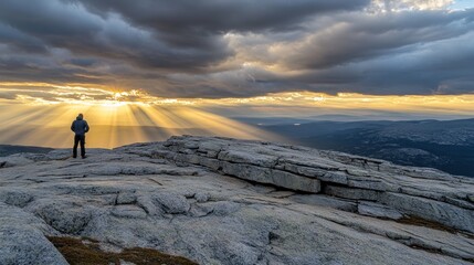 Wall Mural - A solitary figure stands on rocky terrain, illuminated by dramatic rays of sunlight at sunset.