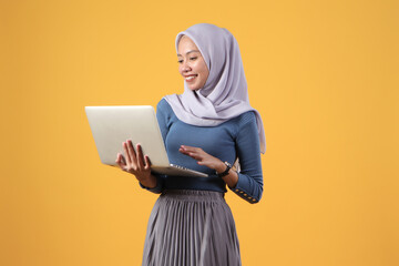 Wall Mural - happy asian indonesian muslim woman holding laptop computer on isolated yellow background