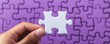 © praewpailyn - A close-up of a person's hand holding a single white puzzle piece against a purple jigsaw puzzle background, symbolizing problem-solving and completion.