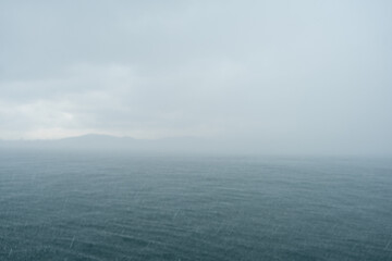 Poster - Light rain creates ripples on the surface of the sea while an overcast sky looms over a tranquil coastal area in the morning light