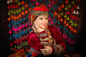 A woman in a colorful headscarf and jewelry poses in front of a wall of colorful tassels