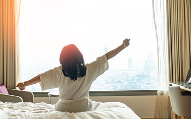 Wall Mural - Hotel relaxation on lazy day with Asian woman waking up from good sleep on bed in weekend morning resting in comfort bedroom looking toward city view, having happy, work-life quality balance lifestyle