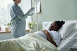 © pressmaster - Medical professional providing care to bedridden patient in hospital room with bright window lighting. Patient lying in bed with attentive healthcare worker standing nearby