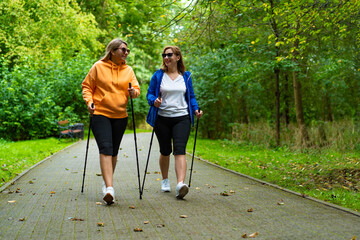 Wall Mural - Two pretty mid-adult women dressed sporty practicing Nordic walking on background of beautiful green trees in city park on spring day. Training of Nordic walking in park.