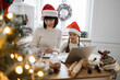 © sofiko14 - Caucasian mother and young daughter wearing Santa hats baking cookies together in cozy kitchen. Festive atmosphere enhanced by Christmas decorations. Focused child working on laptop as mother writes.