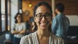 © VK Studio - A woman with glasses smiles warmly in a bustling office environment, exuding positivity and approachability among colleagues.