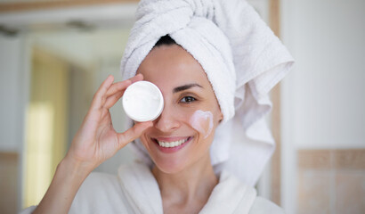 Wall Mural - A smiling woman in a bathrobe and towel holds a cream jar over one eye while a heart-shaped lotion spot is on her cheek, suggesting playful skincare. Perfect for beauty, self-care, or cosmetic ads.