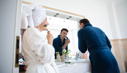 Wall Mural - A man in a blue bathrobe shaves in front of a bathroom mirror while a woman in a white bathrobe, with a towel on her head, uses a face roller. The couple enjoys a shared morning skincare routine.