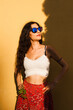 © Nikola Spasenoski - Fashionable young woman with curly hair in sunglasses posing confidently against a vibrant yellow wall on a bright summer day.