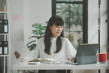 Young asian accountant is working at her desk, checking financial documents and using her tablet