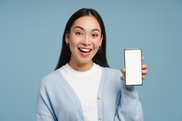 Excited asian woman showing smartphone with blank screen, standing isolated on blue background