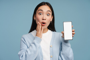Shocked young woman showing smartphone with empty screen touching face with hand, looking camera