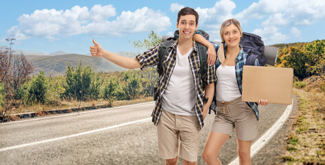 Wall Mural - Cheerful young couple hitchhiking and holding a blank cardboard sign