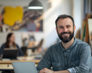 Portrait of cheerful and creative bearded architect looking at camera in modern coworking environment of design studio near colleagues with laptop on blurred background, banner  