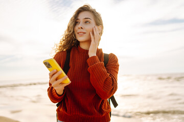 Wall Mural - Young woman smiling and using her smartphone at the beach during sunset, with golden sunlight illuminating her hair and surroundings. Blogging, communication concept.