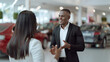 © tikhomirovsergey - Smiling african american salesman is talking to the customer at car showroom. Professional salesperson working in modern car dealership. Modern technology in auto dealership. Modern transportation.