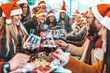 © Davide Angelini - Happy people wearing santa claus hat having Christmas dinner party - Cheerful group of friends celebrating new year together - Winter holidays concept