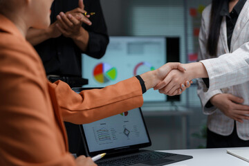 Two businesswomen shaking hands during a work meeting with their manager applauding in the background near a whiteboard