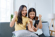 © amnaj - Two women celebrating on a couch, looking at a smartphone in a bright living room, sharing a moment of success and connection