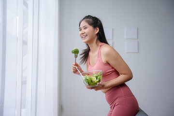 Wall Mural - Young woman is smiling while eating a healthy salad at home after exercising wearing sportswear