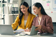 © amnaj - Two young businesswomen are having a working discussion while sitting at an office desk and using a laptop computer