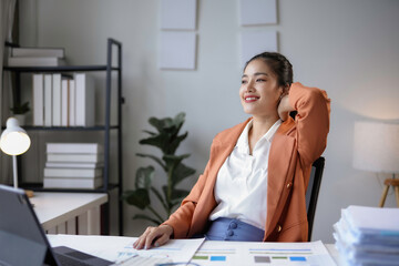 Poster - Young asian businesswoman is leaning back in her chair with her hand behind her head, smiling and looking away from the camera