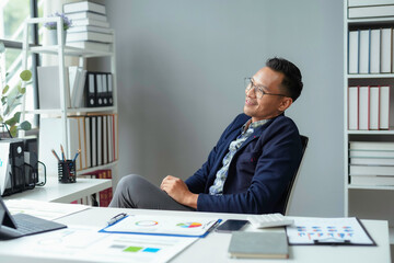 Poster - Asian businessman is sitting at his desk relaxing and smiling after finishing work in a modern office