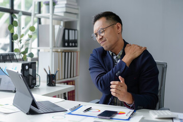 Young businessman is holding his shoulder in pain while working at his desk in a bright office