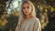 © Sadia - Young woman in a casual beige sweater stands in a sunlit forest during autumn