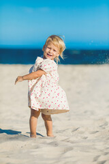 2 years old girl in white clothes sits on the white sand on the beach in summer