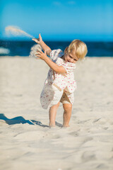 2 years old girl in white clothes sits on the white sand on the beach in summer