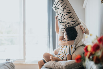 Wall Mural - Two friends are enjoying free time together, playfully engaging in a pillow fight in a sunlit living room. The scene captures a joyful and relaxed atmosphere.