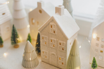 Wall Mural - Merry Christmas! Stylish Christmas miniature holiday village with white houses, trees and golden lights bokeh on snowy window. Atmospheric magical scene. Christmas holiday banner.