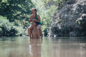 Wall Mural - A joyful couple having fun in a tranquil river environment. The woman rides on the man's shoulders as they splash in the water, surrounded by natural beauty.