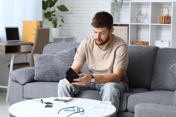 Wall Mural - Young man in debt with empty wallet sitting on sofa at home