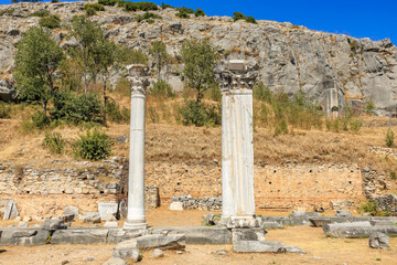 Two white pillars stand in a field next to a rocky hill. Ruins of the ancient city of Philippi, Greece