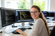 © SejutaCahaya - A smiling young woman with glasses sits at her desk in front of two computer monitors, working on code.