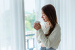 © Monster Ztudio - Young asian woman standing beside window and holding mug in bedroom at home, She drinking milk after wake up in the morning