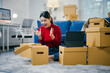 © Parichat - Young woman is sitting on the floor surrounded by cardboard boxes and is unpacking her belongings in her new apartment. She is excited and is having a video call on her tablet