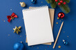 © ActionGP - A top view of a blank notebook surrounded by festive Christmas decorations, including ornaments and a pencil, on a blue background
