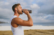 © ViDi Studio - Sideways young strong athletic toned fit sportsman man in sports clothes earphones drink protein shake warm up train at sunrise sun dawn over sea beach outdoor on pier seaside in summer day morning.
