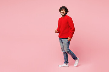 Full body young side view young cheerful happy joyful Indian man wear red sweater casual clothes walk go look camera isolated on plain pastel light pink background studio portrait. Lifestyle concept.