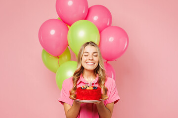 Young woman wears shirt celebrating birthday holiday and hold bunch of colorful air balloons, red cake making wish blow out candles isolated on plain pastel light pink background. Lifestyle concept.