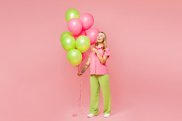 Wall Mural - Full body young woman wears shirt celebrating birthday holiday stand hold bunch of colorful air balloons point index finger aside on area isolated on plain pastel pink background. Lifestyle concept.