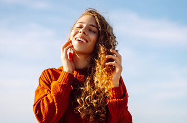 Wall Mural - Young woman with curly hair enjoys the sunny beach, smiling and basking in the warmth of a clear day. Spring time. Travel, weekend, relax and lifestyle concept.